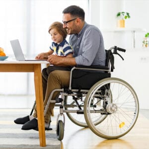 Child sitting on fathers lap at the table with a laptop in front of them. The man is in a wheelchair